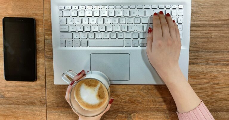 Hands typing on a laptop next to a cappuccino and a smartphone on a wooden table during an AI job search in Germany