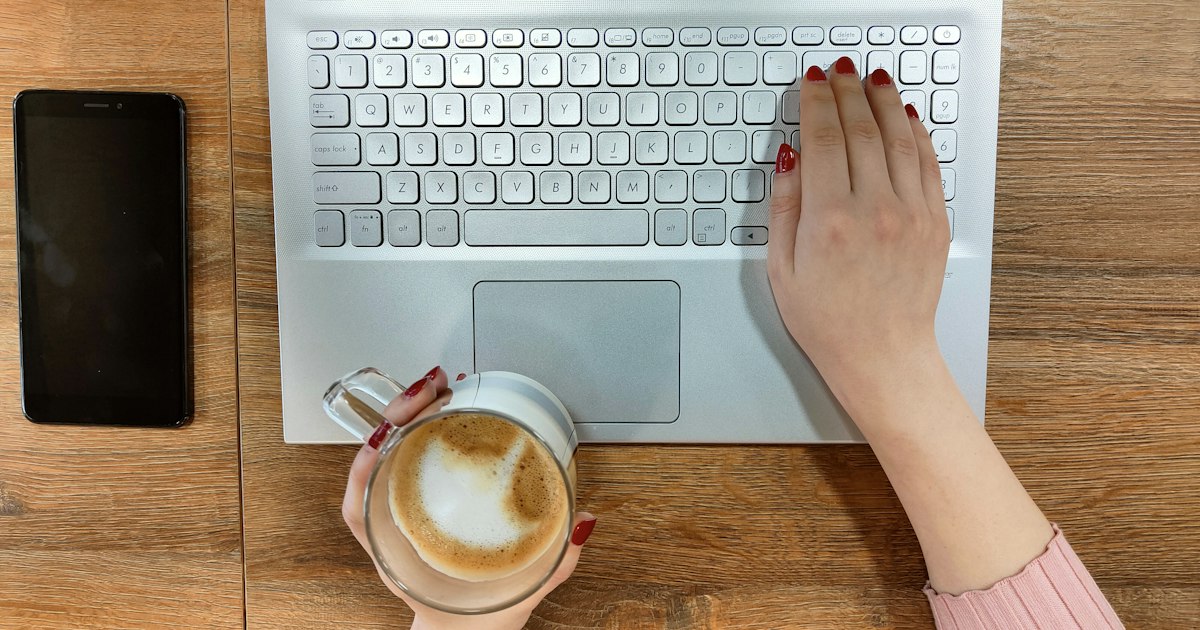 Hands typing on a laptop next to a cappuccino and a smartphone on a wooden table during an AI job search in Germany