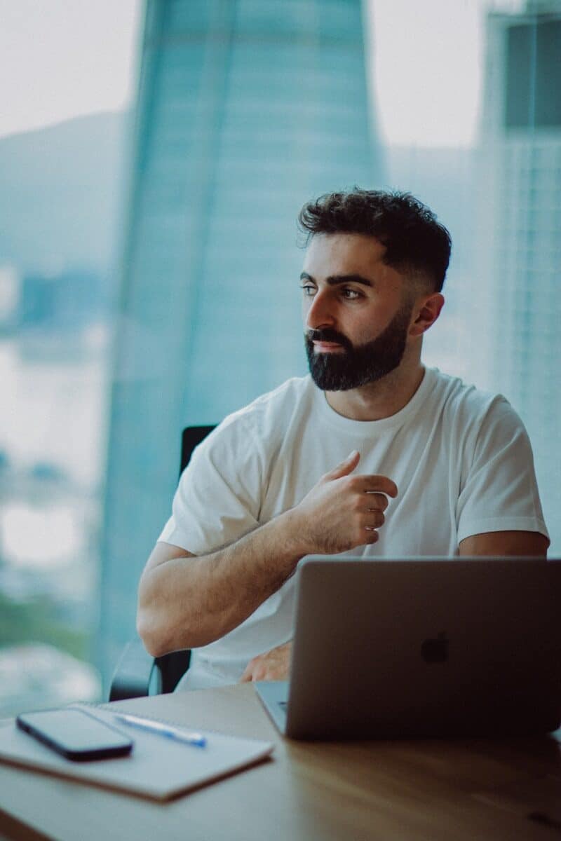 Man with beard working on laptop by window.